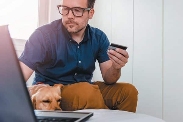 Man making a purchase on his laptop with a dog resting his head on his lap.
