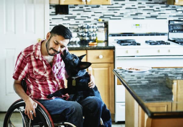 Smiling man in wheelchair having face licked by dog while hanging out in kitchen