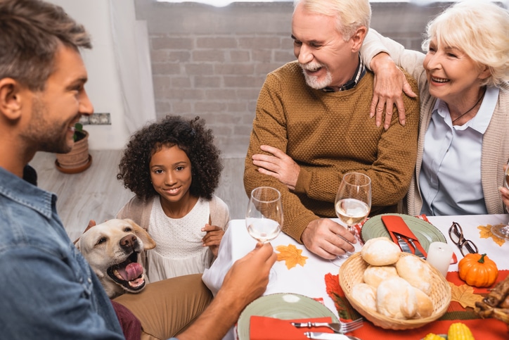 joyful multicultural family holding glasses of white wine while celebrating thanksgiving near golden retriever