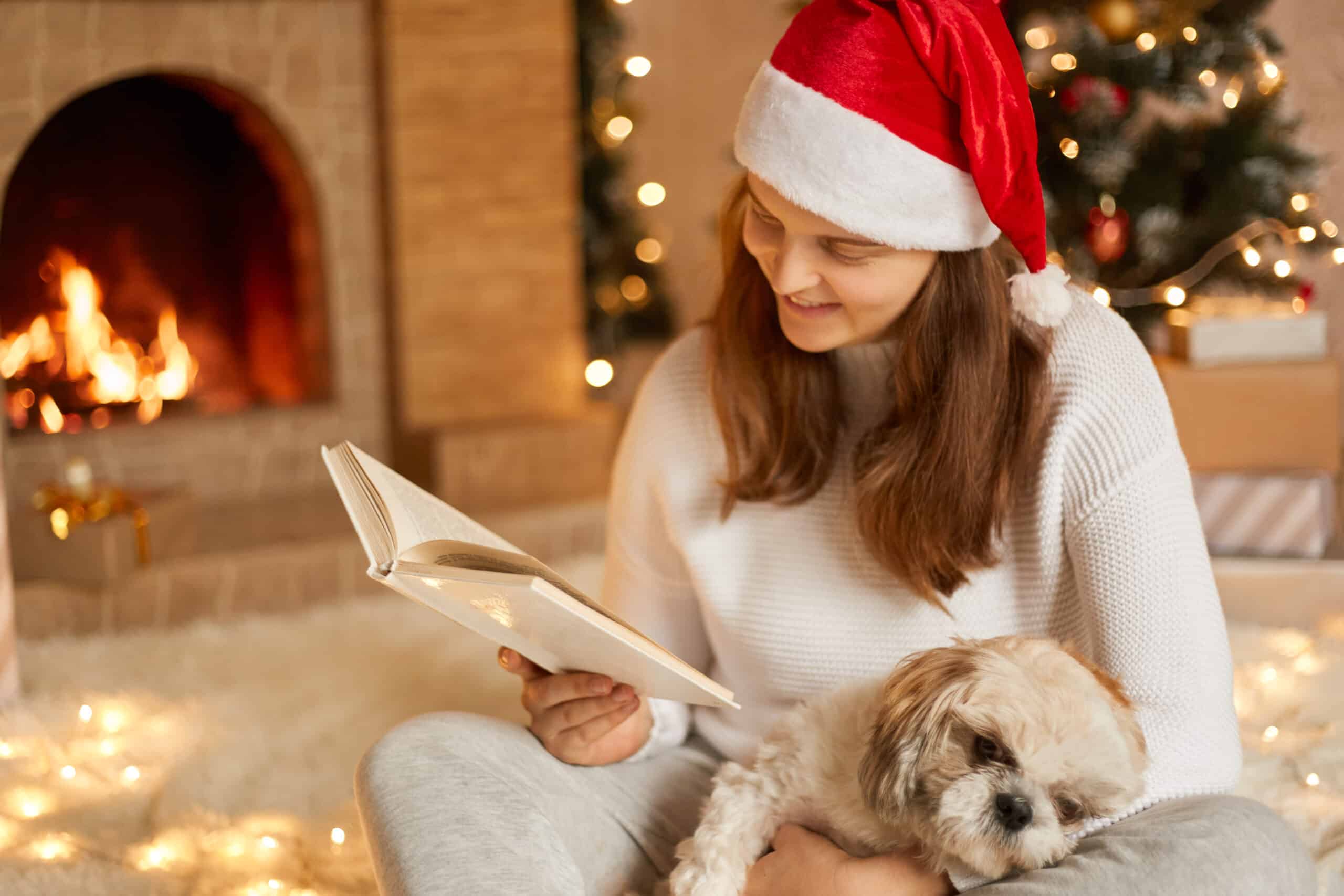 Young woman at home reading book with dog on her knees, looking at pages, wearing casual attire and red festive hat, posing in living room with fireplace and xmas tree on background.