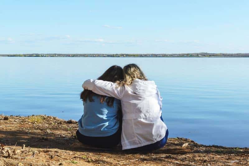 view from the back of two people who are sitting on the shore near the water close to each other hugging