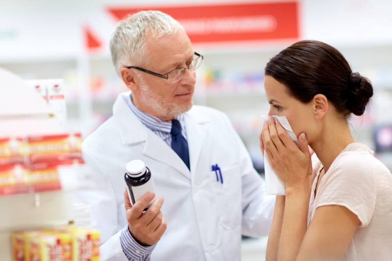 senior apothecary with drug and female customer having cold and blowing nose at pharmacy