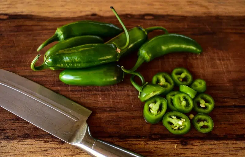 sliced and whole jalapeno peppers on wooden cutting board beside stainless knife