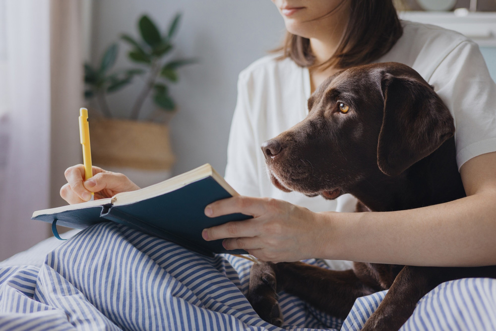 woman writing on notebook with her dog on bed