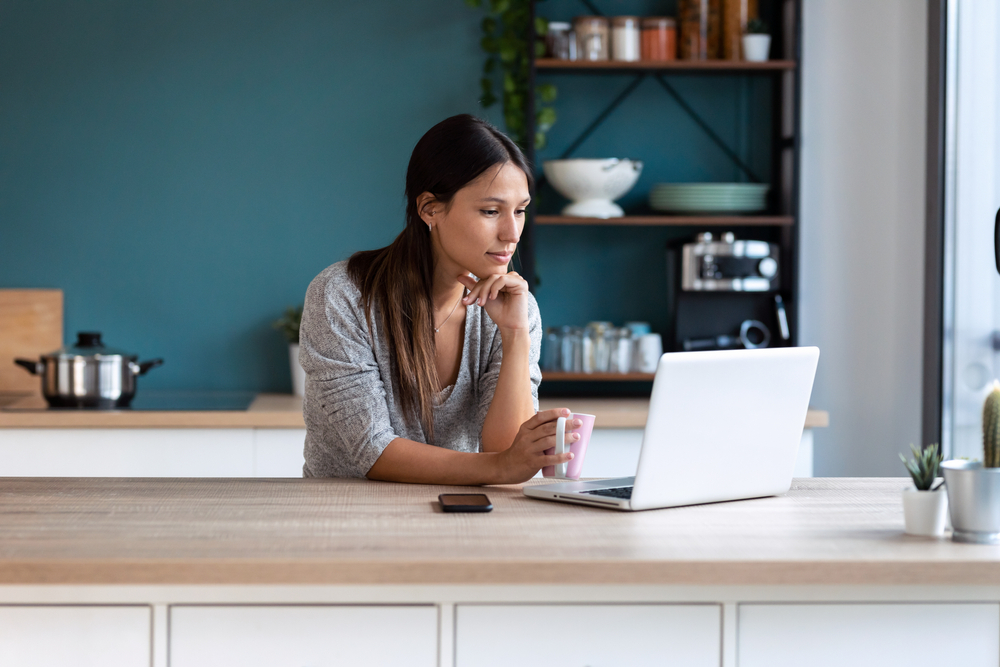 young woman looking her laptop while holding a cup of coffee in the kitchen