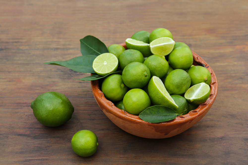 Whole and cut fresh ripe limes with green leaves in bowl on wooden table